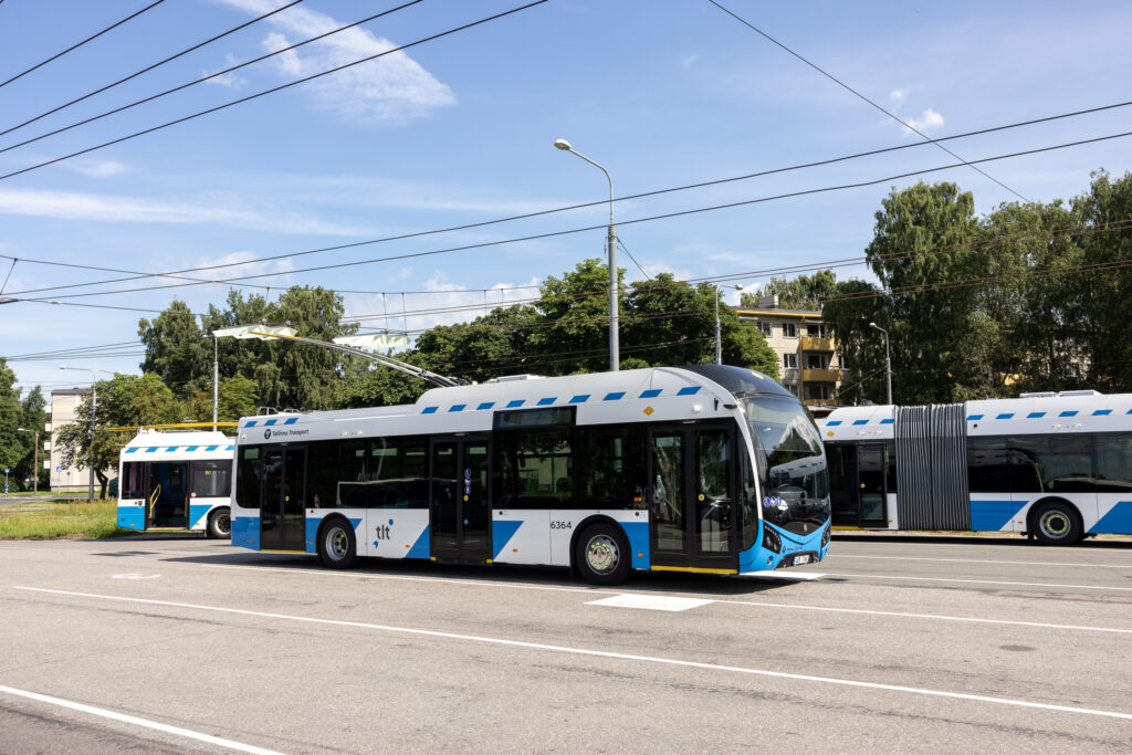 Modernsed sinivalged trollibussid seisavad tänaval päikesepaistelise ilmaga, taustal elektriliinid, puud ja hooned.