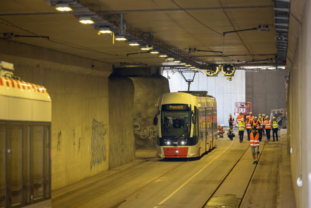 Valge-punane tramm sõidab tunnelis, kus helkurvestides töötajad rööbaste ääres tööd kontrollivad.