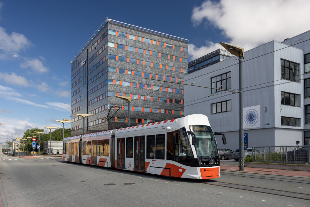 Kaasaegne valge-oranž tramm sõidab linnatänaval moodsate hoonete ees päikeselisel päeval.