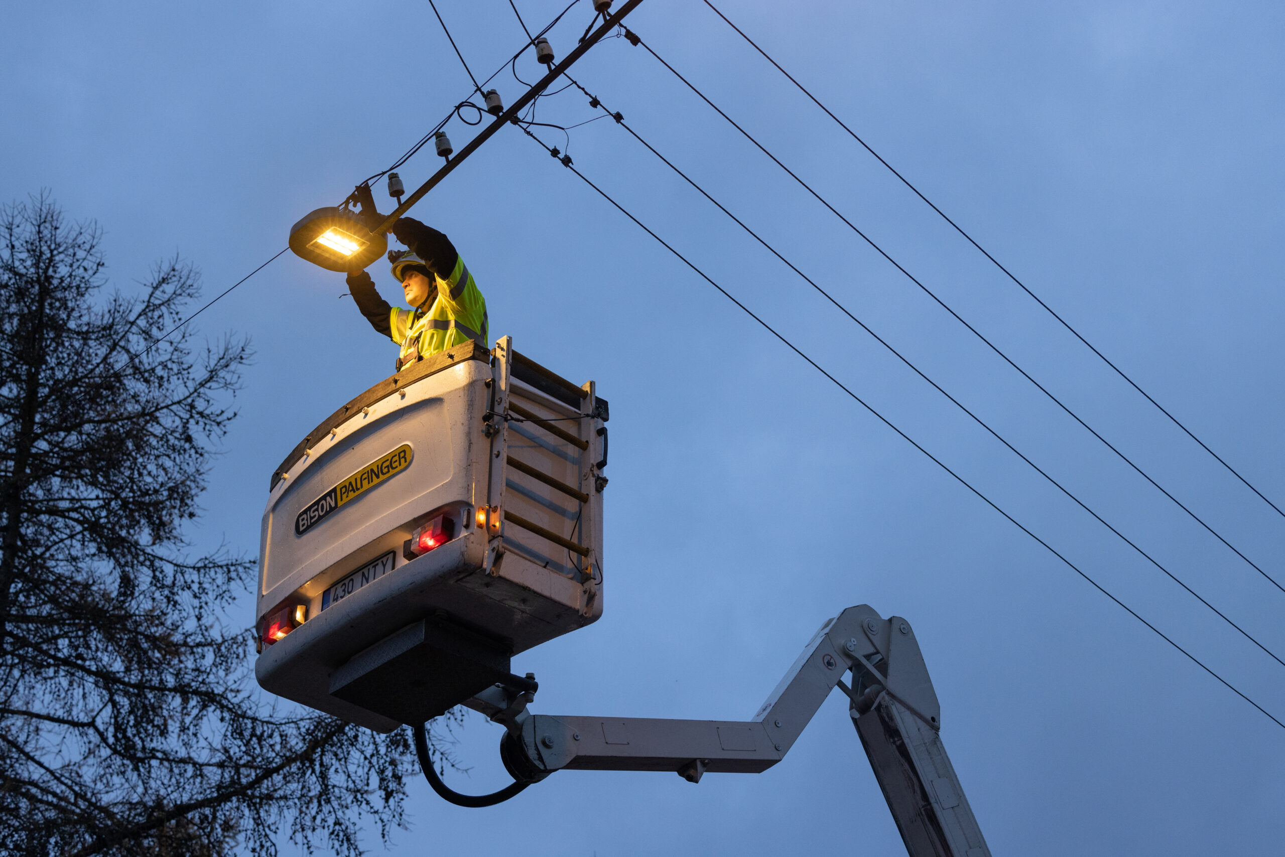 Töömees paigaldab tõstukiga helendavat tänavavalgustit elektriliinide all pimedas taevas.