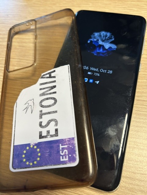 Must nutitelefon ekraaniga, mille kõrval on läbipaistev ümbris Eesti numbrimärgi kleepsuga.